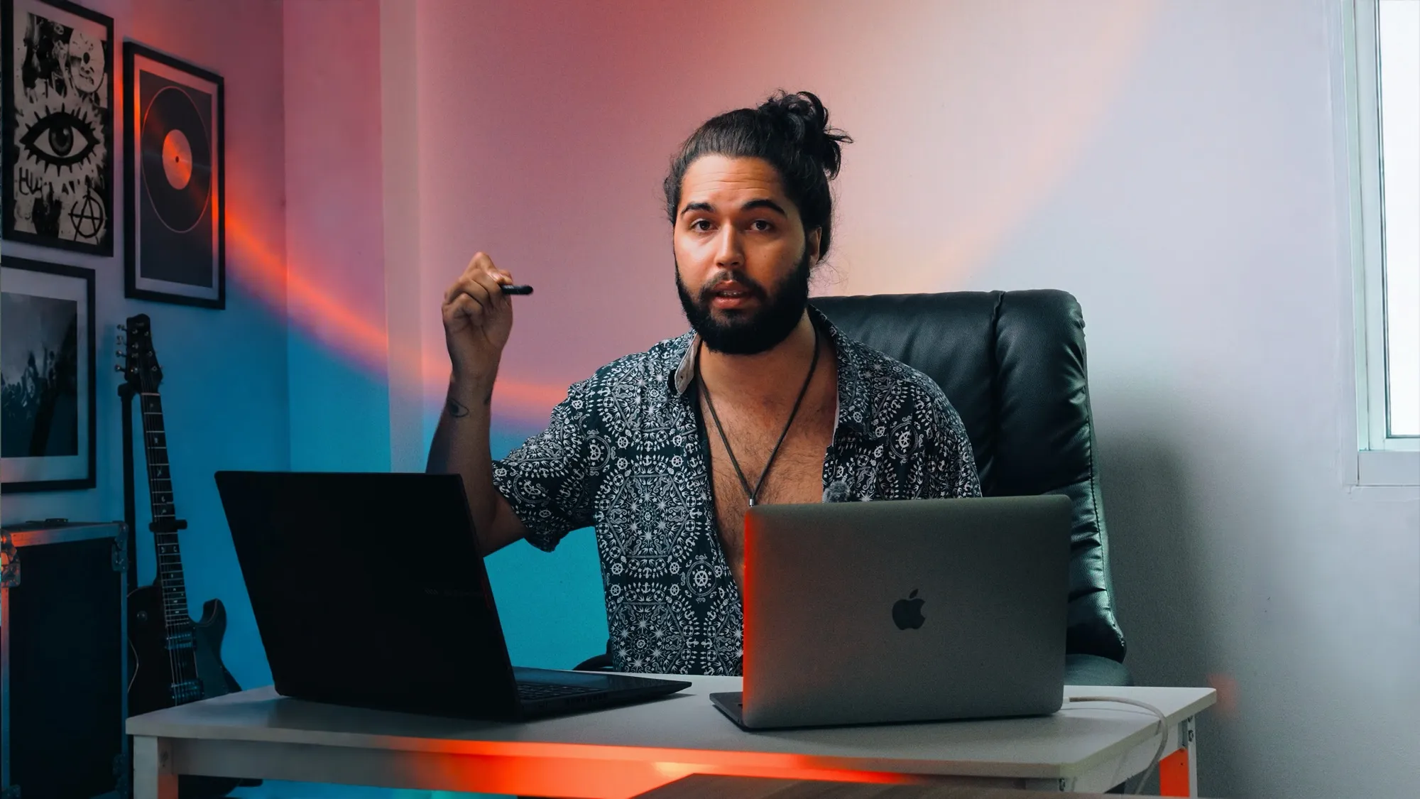 A musician at a desk with two laptops, a guitar and framed posters behind him